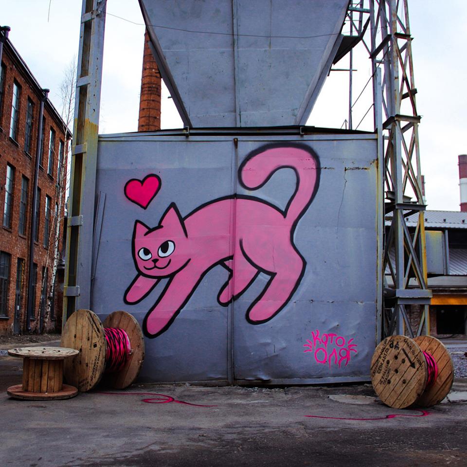 View of industrial area with a pink cat spraypainted on a wall with large wooden spools of wire in front so as to indicate that the cat may be playing with their string